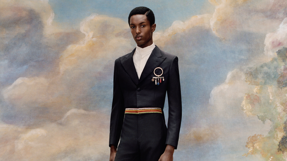 An African American man stands against a cloudy-sky background, wearing a tailored dark suit with medals and a striped belt.